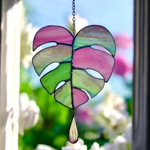 May include: A stained glass suncatcher in the shape of a monstera leaf, with pink, green, and clear glass panels. The suncatcher hangs from a chain and features a teardrop-shaped glass pendant at the bottom. The background is a blurred view of a window and flowers.