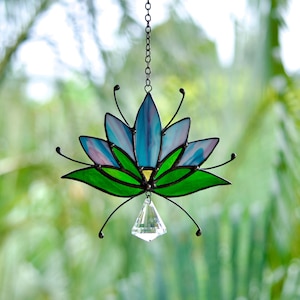 May include: Stained glass lotus flower suncatcher with blue, green, and pink hues. The lotus has a clear crystal pendant hanging from the center. The suncatcher is suspended by a black chain.