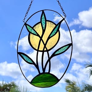 May include: Stained glass art piece featuring a stylized plant design. The artwork includes a yellow sun, green leaves, and a green base, all framed within a black oval. A chain is attached for hanging. The background shows a blue sky with clouds.
