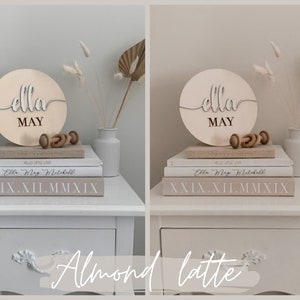 May include: A wooden sign with the name "Ella May" in cursive script, displayed on a stack of books with a small wooden toy and a white vase with dried flowers. The sign is on a white nightstand with a drawer.