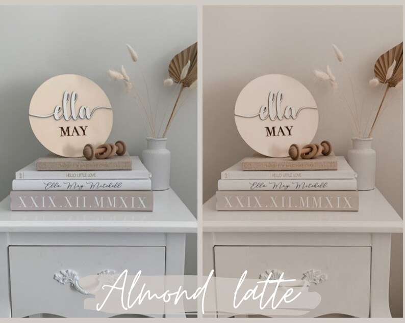 May include: A wooden sign with the name "Ella May" in cursive script, displayed on a stack of books with a small wooden toy and a white vase with dried flowers. The sign is on a white nightstand with a drawer.