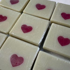 May include: A collection of square, off-white soaps, each featuring a vibrant red heart in the center. The soaps are individually wrapped in clear plastic, creating a clean and appealing presentation. Ideal for gifts or personal use.