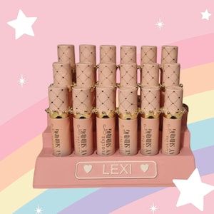 Pode incluir: Exibição de batons rosa com tampas douradas em forma de coroa. Os tubos estão dispostos em um expositor rosa com o nome "LEXI" na frente. Fundo de arco-íris pastel e estrelas.