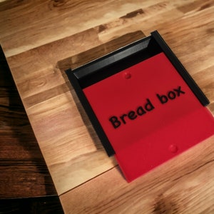 May include: A black and red bread box with the words "Bread box" printed in black on the red front panel.