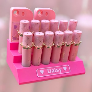May include: A pink lipstick display featuring multiple tubes of lipstick. The lipsticks are in a pink and gold color scheme, with a crown design on the cap. The display has the word "Daisy" on it.