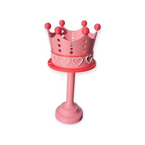May include: A pink crown-shaped holder with a round base and a long stem. The crown has heart and diamond cutouts, and is topped with red spheres. The holder is designed to hold a small object.