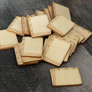 May include: A pile of small, square wooden tiles with a light brown finish. Each tile features a rectangular border with etched lines and star-shaped accents. The tiles are stacked and scattered on a dark grey wooden surface.