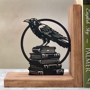 May include: Black metal bookends featuring a raven perched on a stack of books. The bookends are on a wooden base and are designed to hold books upright.