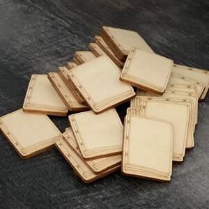 May include: A pile of small, square wooden book-shaped cutouts. Each has a light-coloured centre with a darker, detailed border. The cutouts are scattered on a dark, textured surface, suggesting a craft or DIY project.
