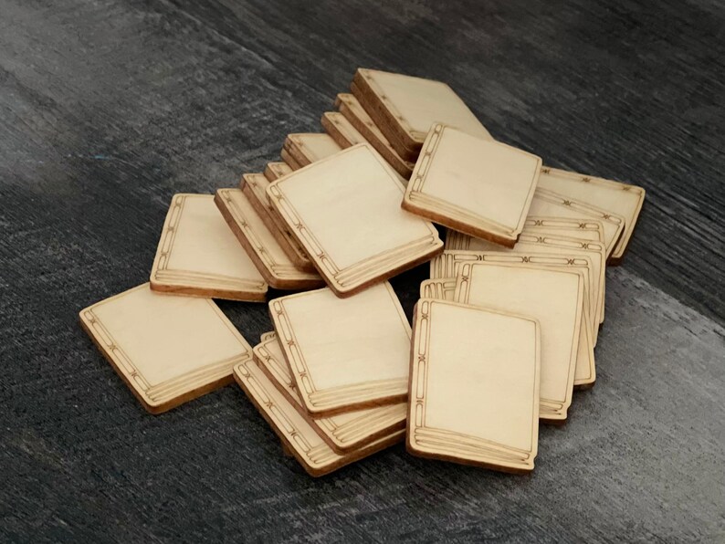 May include: A pile of small, square wooden book-shaped cutouts. Each has a light-coloured centre with a darker, detailed border. The cutouts are scattered on a dark, textured surface, suggesting a craft or DIY project.