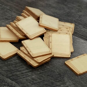 May include: A pile of unfinished, square wooden game pieces with rounded corners. Each piece has a light wood grain surface and a decorative border. The pieces are scattered on a dark grey surface.