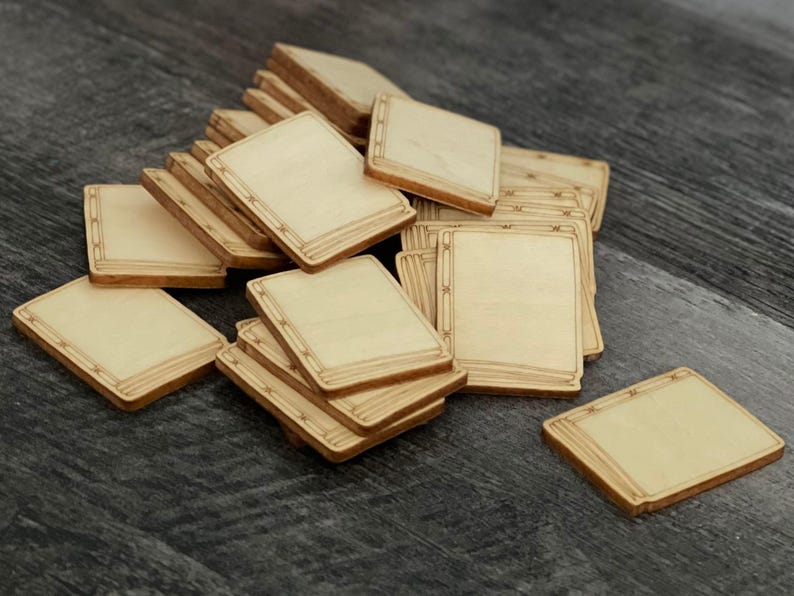 May include: A pile of unfinished, square wooden game pieces with rounded corners. Each piece has a light wood grain surface and a decorative border. The pieces are scattered on a dark grey surface.