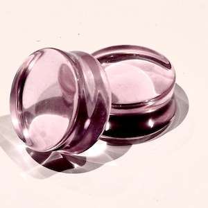 May include: Two clear, light purple ear plugs. The plugs are circular with a flat front and back, and a slightly raised rim. The plugs are made of a shiny, transparent material, and cast a shadow on the white surface.