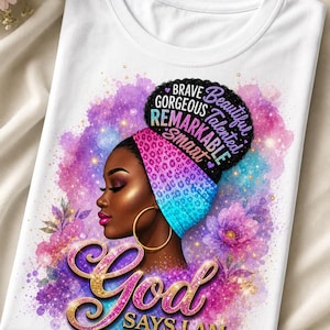 Peut inclure: T-shirt blanc avec une illustration vibrante d'une femme noire portant un foulard coloré et des créoles dorées. Le design comprend les mots "Dieu dit que je suis" et d'autres affirmations positives.