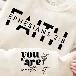 Puede incluir: Una sudadera de color crema con texto negro que dice "FAITH Ephesians 2:8" y "you are worth it" con un pequeño diseño floral.