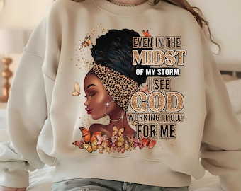 May include: Cream-colored sweatshirt featuring a graphic of a Black woman with a leopard print head wrap, surrounded by butterflies. The text reads "Even in the Midst of My Storm I See God Working It Out For Me."