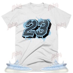 K&ouml;nnte beinhalten: Ein wei&szlig;es T-Shirt mit einer schwarzen und blauen Grafik der Nummer 23. Die Nummer tropft mit blauer Farbe. Das T-Shirt wird mit einem Paar hellblauer Sneakers mit wei&szlig;er Sohle kombiniert.