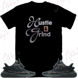 May include: Black t-shirt with the text "Hustle & Grind" in a gray and brown script font. The t-shirt is paired with a pair of black and green camouflage sneakers.