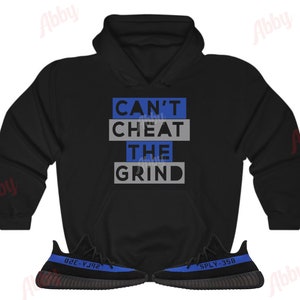 Może przedstawiać: Czarna bluza z kapturem z napisem "Can't Cheat The Grind" w niebieskich i szarych literach. Bluza jest połączona z czarnymi i niebieskimi butami sportowymi z napisem "SPLY-350" z boku.