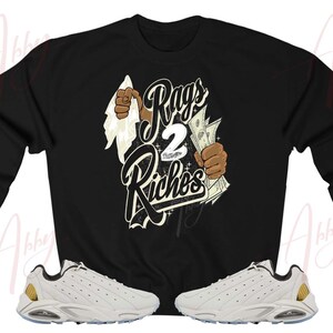 May include: Black sweatshirt with a graphic print of a hand tearing through a white cloth revealing the text "Rags 2 Riches". The sweatshirt is paired with white sneakers with gold accents.