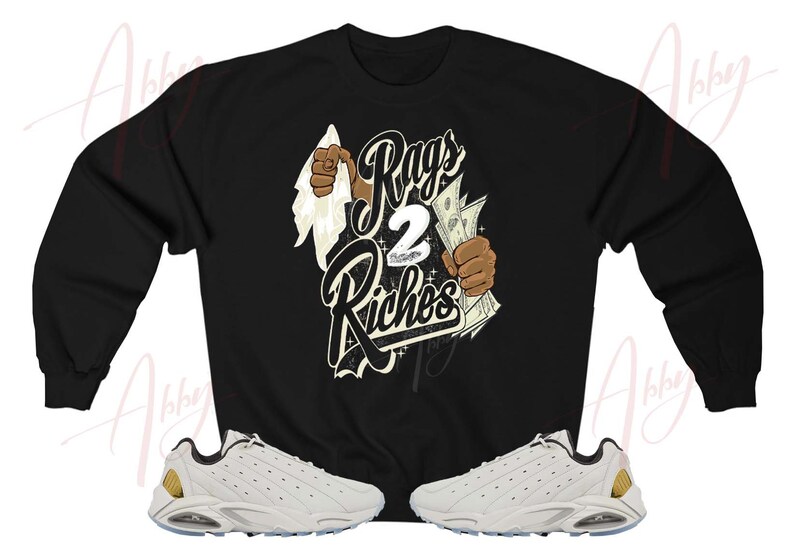 May include: Black sweatshirt with a graphic print of a hand tearing through a white cloth revealing the text "Rags 2 Riches". The sweatshirt is paired with white sneakers with gold accents.