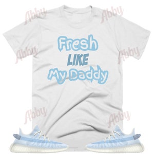 May include: A white t-shirt with the text "Fresh Like My Daddy" in blue lettering. The shirt is paired with a pair of light blue trainers with white soles.