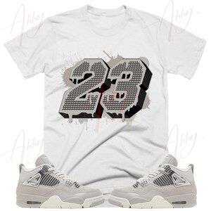 May include: A white t-shirt with a gray and black graphic of the number 23 with paint splatters. A pair of gray and white sneakers with a mesh design.