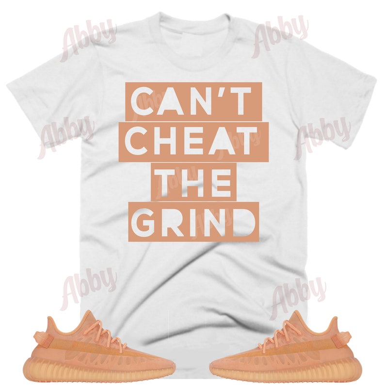 May include: A white t-shirt with the text "Can't Cheat The Grind" in peach colored boxes. Two pairs of peach colored sneakers are on either side of the t-shirt.