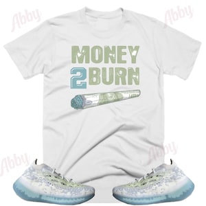 May include: White t-shirt with the text "MONEY 2 BURN" in green and blue lettering. A green and blue rolled-up dollar bill is below the text. A pair of white and blue sneakers with a camouflage pattern are below the t-shirt.
