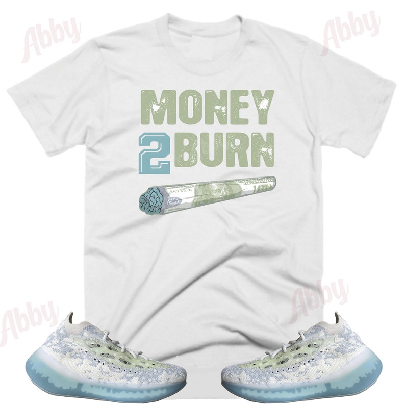 May include: White t-shirt with the text "MONEY 2 BURN" in green and blue lettering. A green and blue rolled-up dollar bill is below the text. A pair of white and blue sneakers with a camouflage pattern are below the t-shirt.
