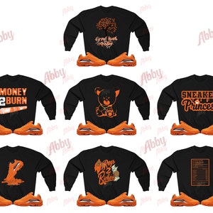 May include: A selection of black sweatshirts featuring orange designs and text, alongside orange trainers. The graphics include phrases such as "Money 2 Burn" and "Sneaker Princess", and illustrations of a bear and a tree.