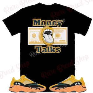 May include: A black t-shirt with a yellow and green graphic of a dollar bill with a mouth and tongue sticking out. The text "Money Talks" is printed below the graphic. Two yellow, black, and orange sneakers are shown below the t-shirt.