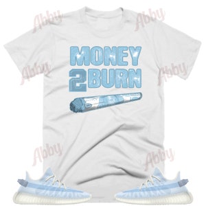 May include: White t-shirt with blue text that says "Money 2 Burn" and a blue image of a rolled-up dollar bill.  The t-shirt is paired with light blue sneakers with white soles.
