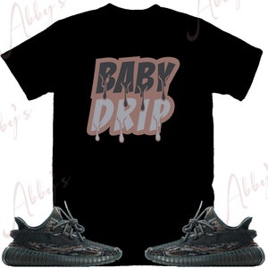 May include: Black t-shirt with the text "BABY DRIP" in pink and black lettering. The shirt is paired with a pair of black and green camouflage sneakers.