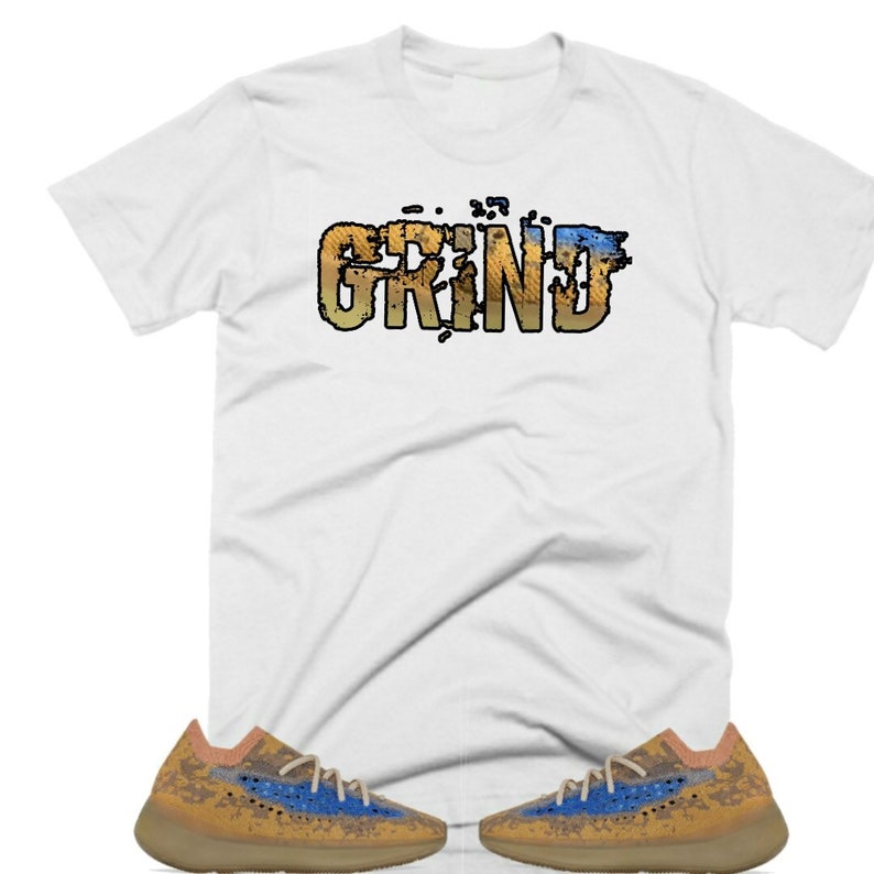 May include: White t-shirt with the word "GRIND" in a distressed, multi-colored font. The shirt is paired with a pair of tan and blue sneakers.