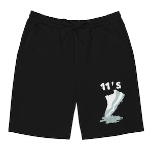 Puede incluir: Pantalones cortos negros con cintura con cordón y bolsillos. Los pantalones cortos presentan un gráfico de una zapatilla deportiva con el texto "11's" en blanco. El diseño de la zapatilla es blanco y azul claro.