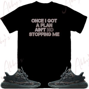 Puede incluir: Camiseta negra con el texto "ONCE I GOT A PLAN AIN'T NO STOPPING ME" en letras blancas y grises. Tambi&eacute;n se muestra un par de zapatillas de deporte de camuflaje negro y verde.