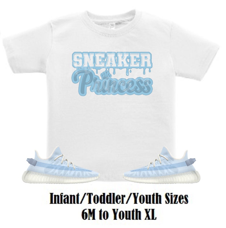 Może przedstawiać: Biała koszulka z niebieskim napisem "Sneaker Princess". Pod koszulką widoczne są dwie jasnoniebieskie i białe buty sportowe. Na dole obrazu znajduje się napis "Infant/Toddler/Youth Sizes 6M to Youth XL".