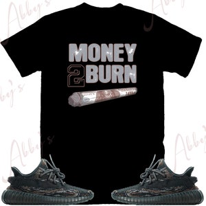 May include: Black t-shirt with the text "MONEY 2 BURN" in white with a silver glitter effect. A rolled-up dollar bill is pictured below the text. A pair of black and green camouflage sneakers are shown below the t-shirt.