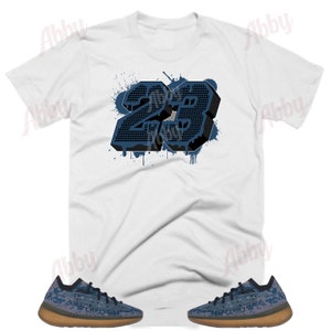 May include: White t-shirt featuring a blue and black number 23 graphic. The number is in a 3D style with a paint splatter effect. The shirt is paired with a pair of blue and brown trainers.
