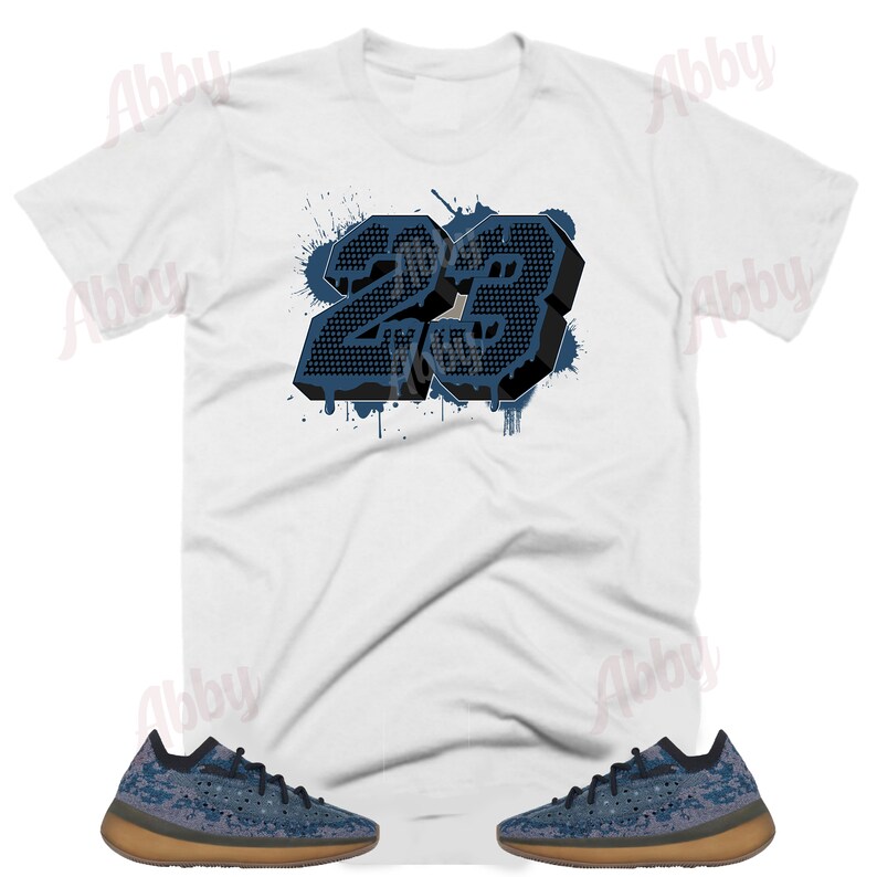 May include: White t-shirt featuring a blue and black number 23 graphic. The number is in a 3D style with a paint splatter effect. The shirt is paired with a pair of blue and brown trainers.