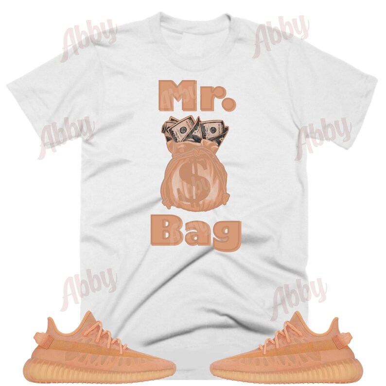 May include: A white t-shirt with the text "Mr. Bag" printed on it in brown. The graphic features a brown money bag with dollar bills spilling out of the top. A pair of light orange sneakers are shown on either side of the t-shirt.