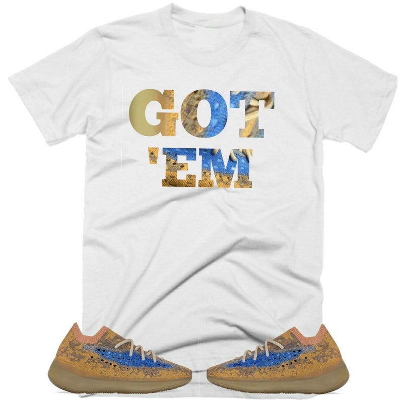 May include: White t-shirt with the text "GOT 'EM" in gold and blue letters. The shirt is paired with a pair of brown and blue sneakers.