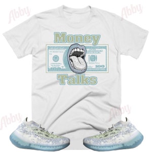 Pode incluir: Camiseta branca com a frase "Money Talks" em verde e um desenho de uma boca e l&iacute;ngua em uma nota de 100 d&oacute;lares. A imagem tamb&eacute;m mostra um par de t&ecirc;nis brancos e azul claro com sola transl&uacute;cida.