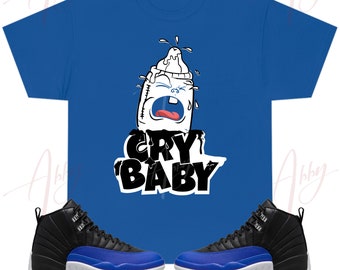 May include: A royal blue t-shirt with a white baby bottle graphic with a crying face and the text "CRY BABY" in black. The t-shirt is paired with black and royal blue sneakers.
