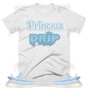 K&ouml;nnte beinhalten: Wei&szlig;es T-Shirt mit blauem Text, der "Princess Drip" mit einer Krone &uuml;ber dem Wort "Drip" sagt. Das Shirt wird mit einem Paar hellblauer und wei&szlig;er Turnschuhe kombiniert.