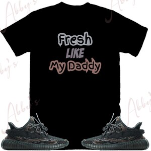 May include: Black t-shirt with the text "Fresh Like My Daddy" in a gray and brown gradient. The t-shirt is paired with a pair of black and green camouflage sneakers.