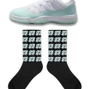 Puede incluir: Una zapatilla deportiva blanca y verde menta con suela blanca se muestra sobre dos calcetines negros. Los calcetines tienen un patrón repetido del número "23" en azul claro.