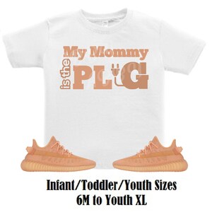 May include: White t-shirt with the text "My Mommy is the PLUG" in peach colored letters. The letter "G" has a plug symbol in it. Two peach colored sneakers are below the shirt. The text "Infant/Toddler/Youth Sizes 6M to Youth XL" is below the sneakers.