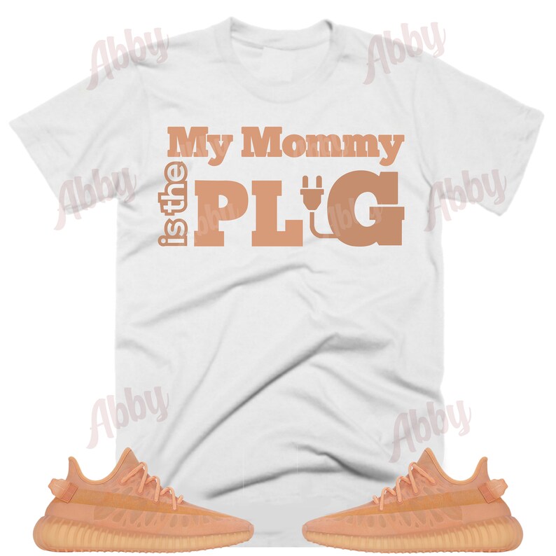 May include: A white t-shirt with the text "My Mommy is the PLUG" in brown with an electrical plug symbol. The shirt is paired with two pairs of light orange sneakers.